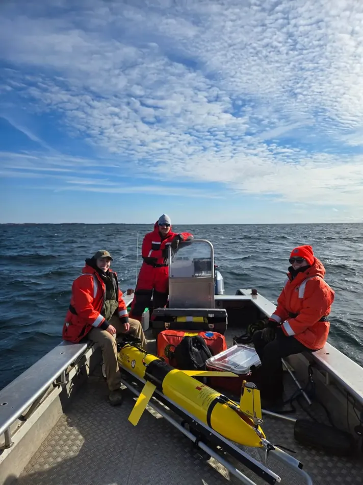 Researchers wearing flotation suits aboard a boat with scientific monitoring equipment.